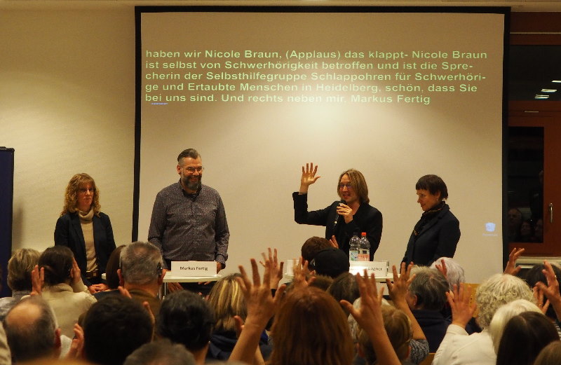 Das Bild zeigt Publikum mit zum Applaus erhobenen Händen und im Hintergrund die Teilnehmer der Podiumsdiskussion
