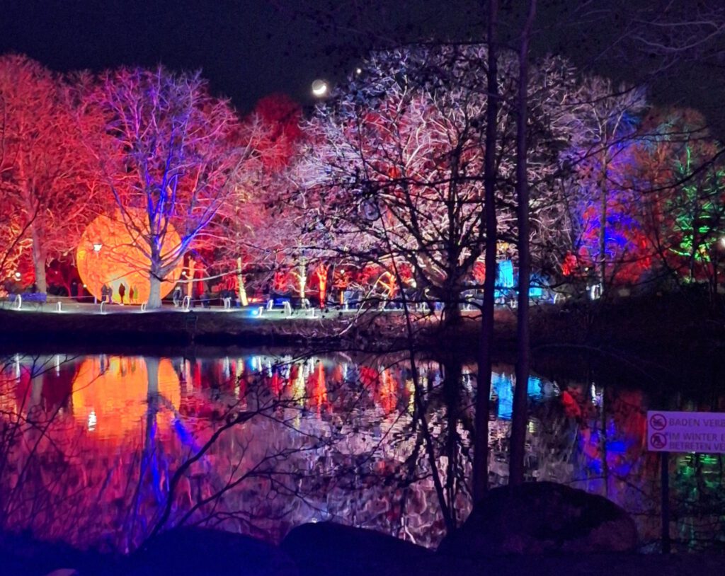 bunt beleuchtete Bäume und ein oranger Globus spiegeln sich im Parksee